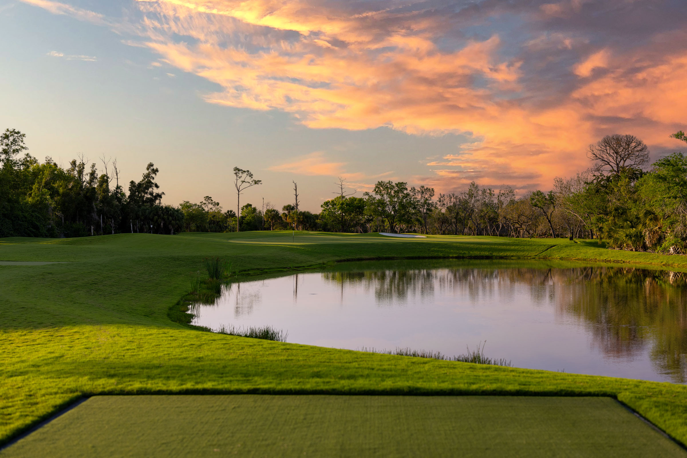 A lush green golf course with a water feature is seen at sunset, under a sky filled with vibrant, orange-hued clouds. Trees line the course in the background.