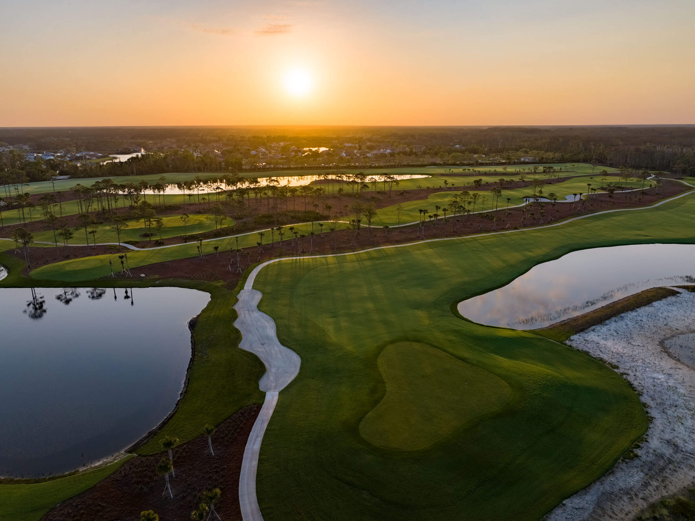 Aerial view of a golf course at sunset, featuring lush greens, water hazards, and a pathway, with a scenic backdrop of a forest and river.