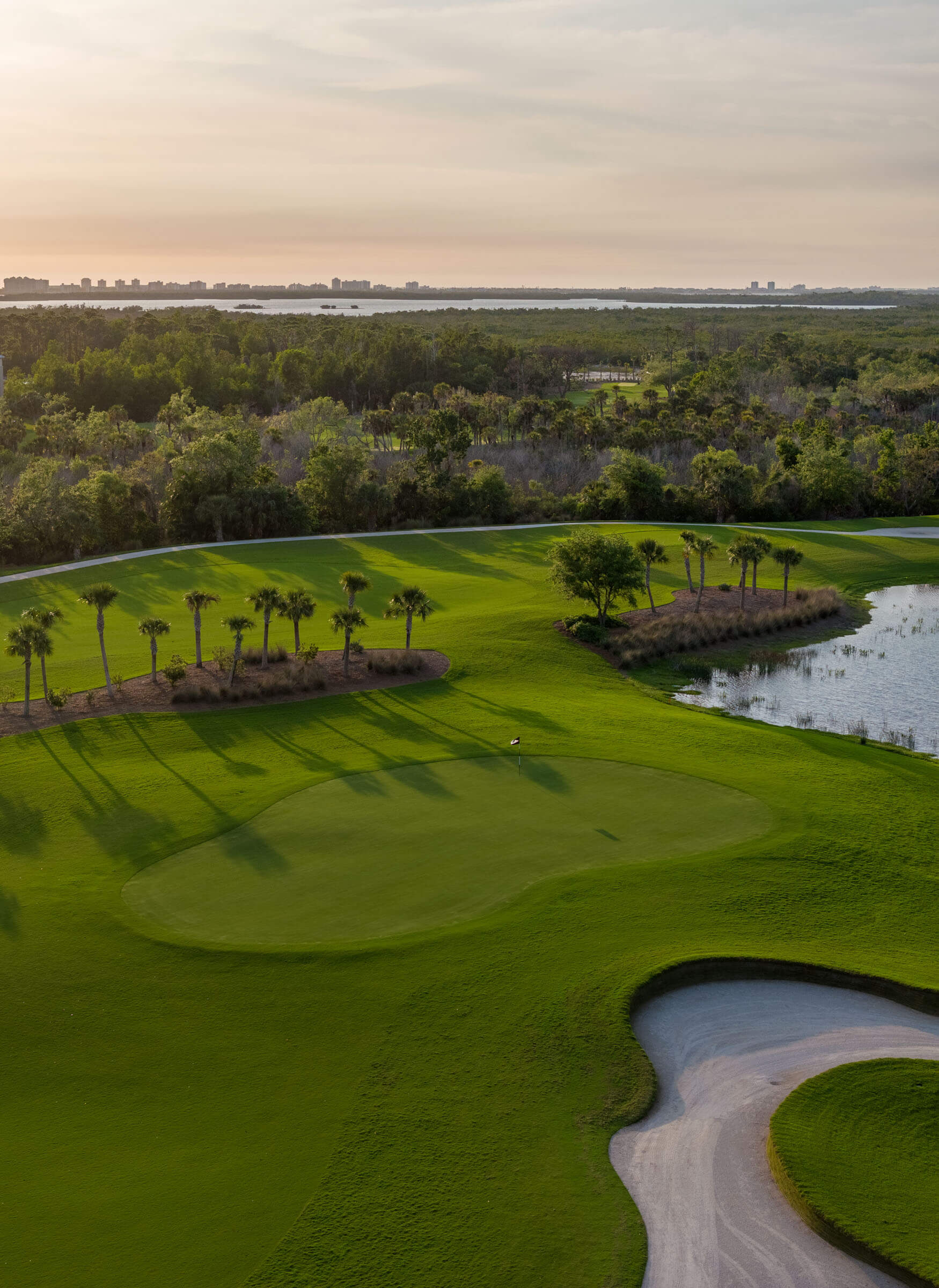Aerial view of a lush golf course with a winding cart path, water hazards, and palm trees during sunset.