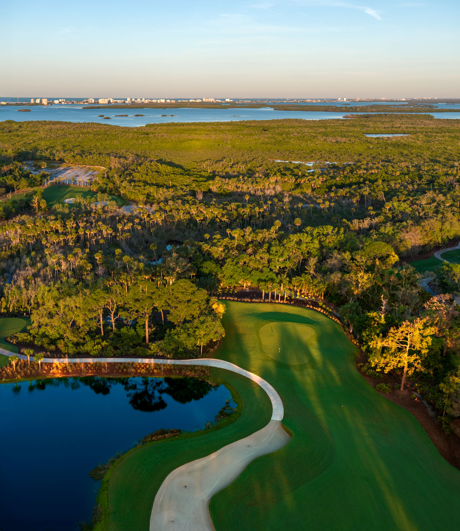 Aerial view of a golf course with a sand trap, water hazard, and neatly trimmed fairway, surrounded by dense greenery and bordered by a body of water in the distance.