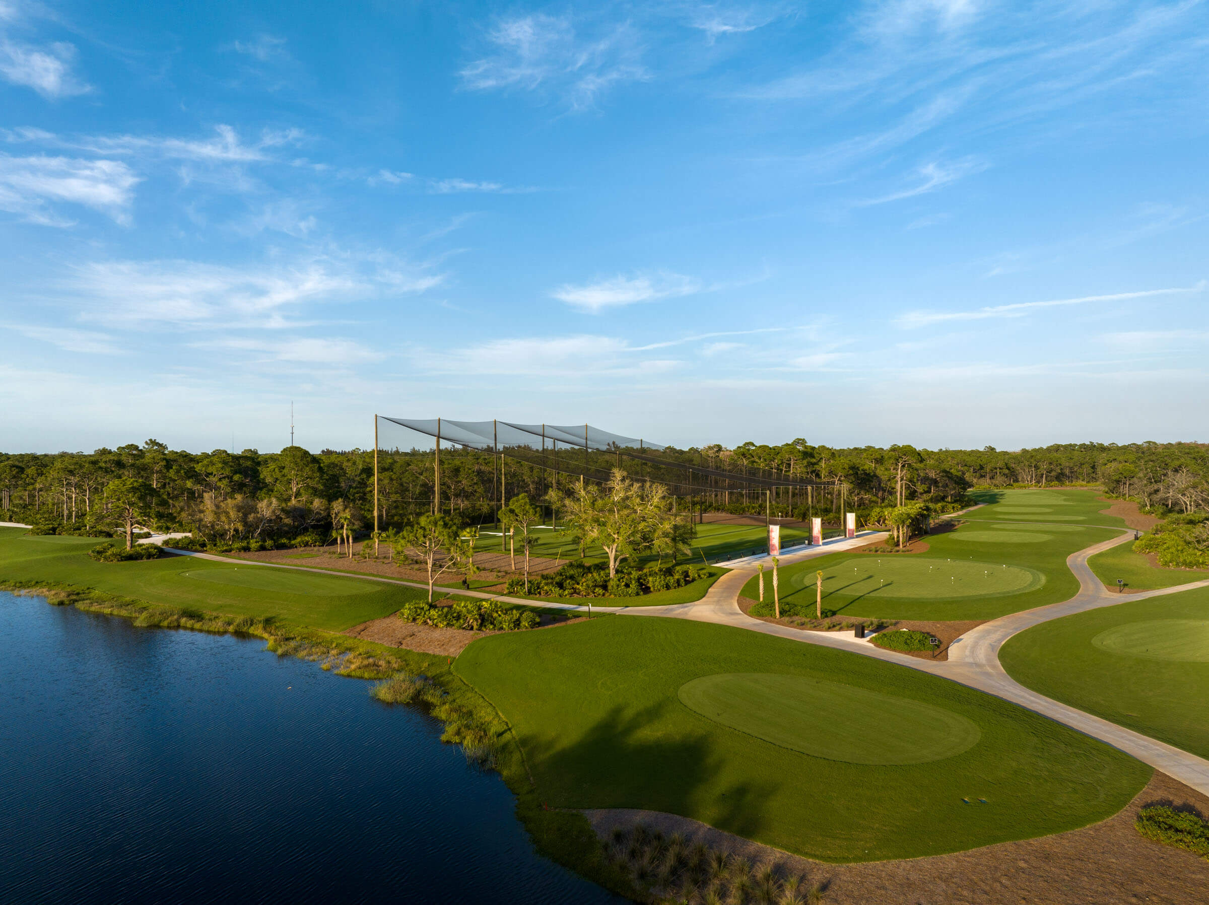 Aerial view of a golf driving range next to a pond with lush greenery and a clear sky.