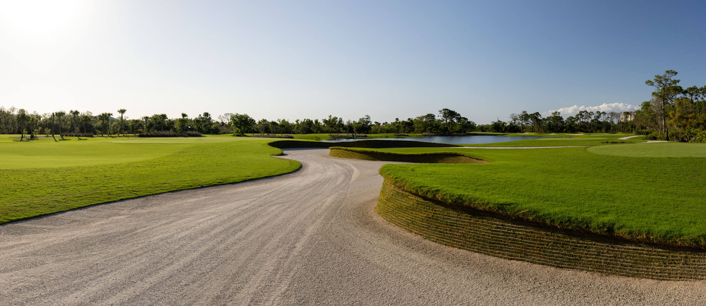 Wide-angle view of a golf course with a curving sand path, manicured green lawns, a water hazard, and scattered clouds in a clear blue sky.