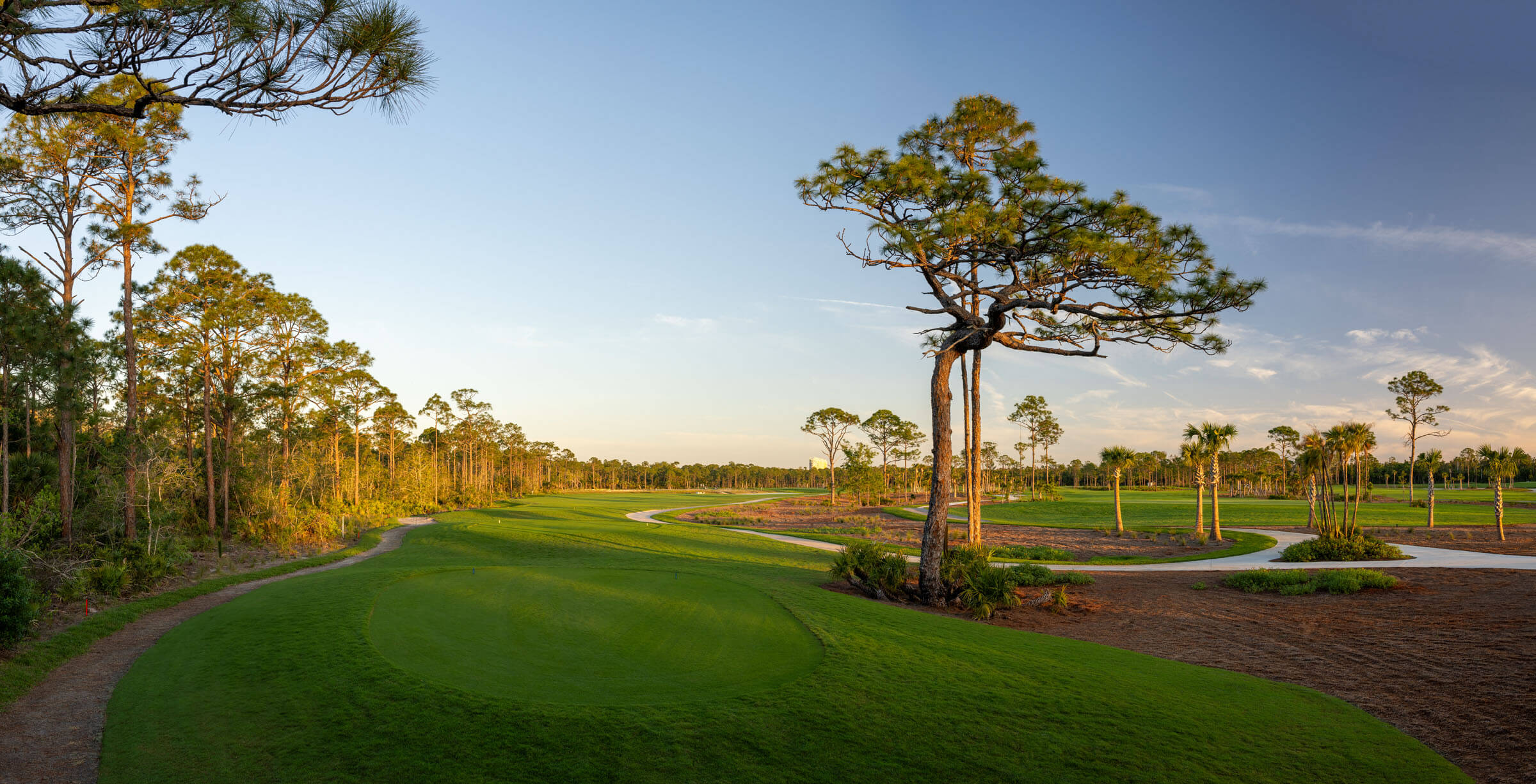 Scenic golf course with lush fairways, pine trees, and a winding path under a clear sky at sunset.