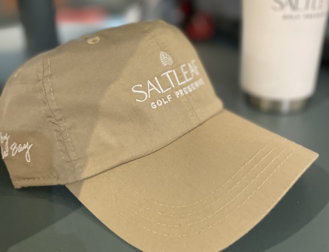 A tan baseball cap with "Saltleaf Golf Preserve" embroidered in white on the front, resting on a surface, with a white tumbler in the background.