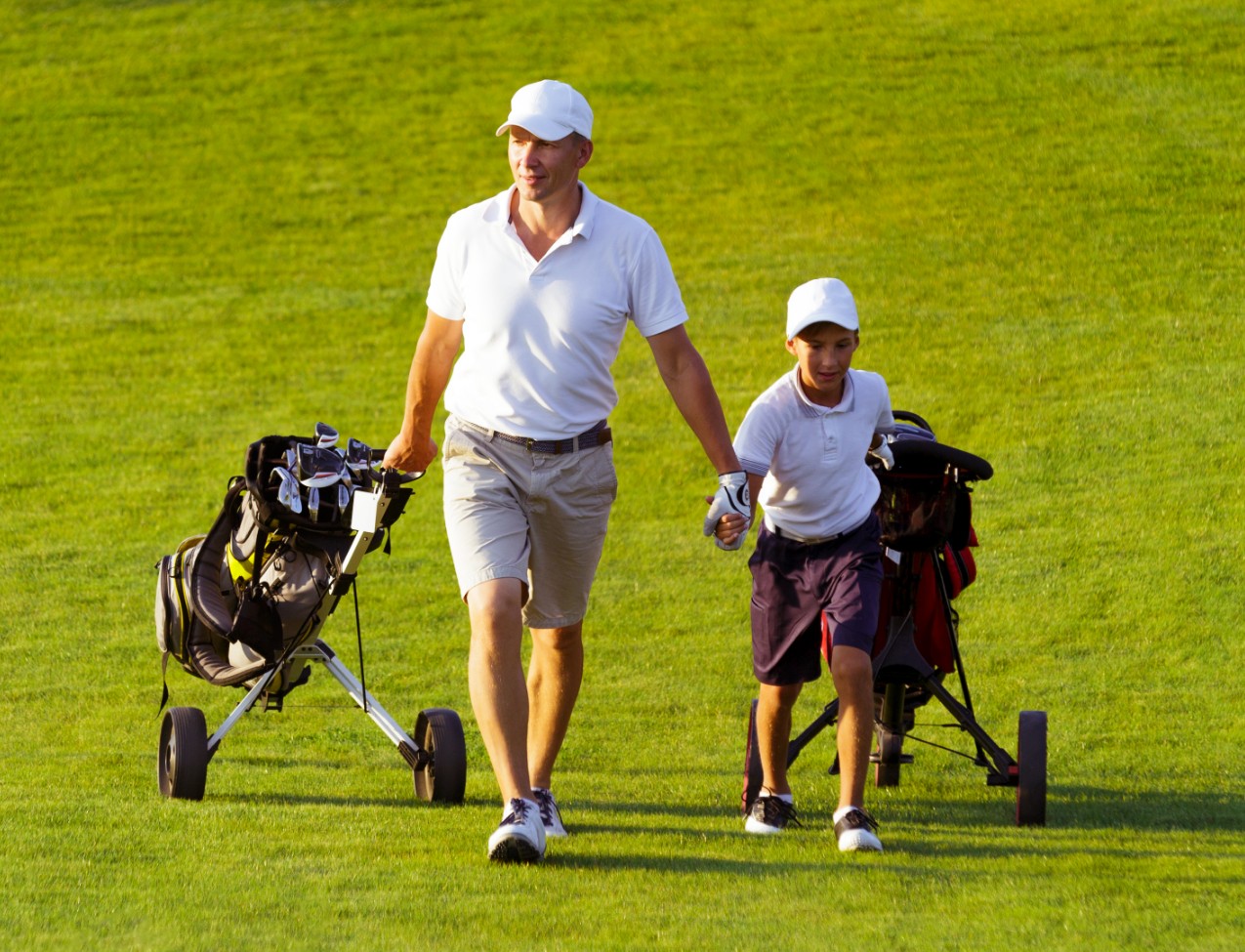 An adult and a child wearing white shirts and hats walk on a golf course, each pulling a golf bag on wheels.