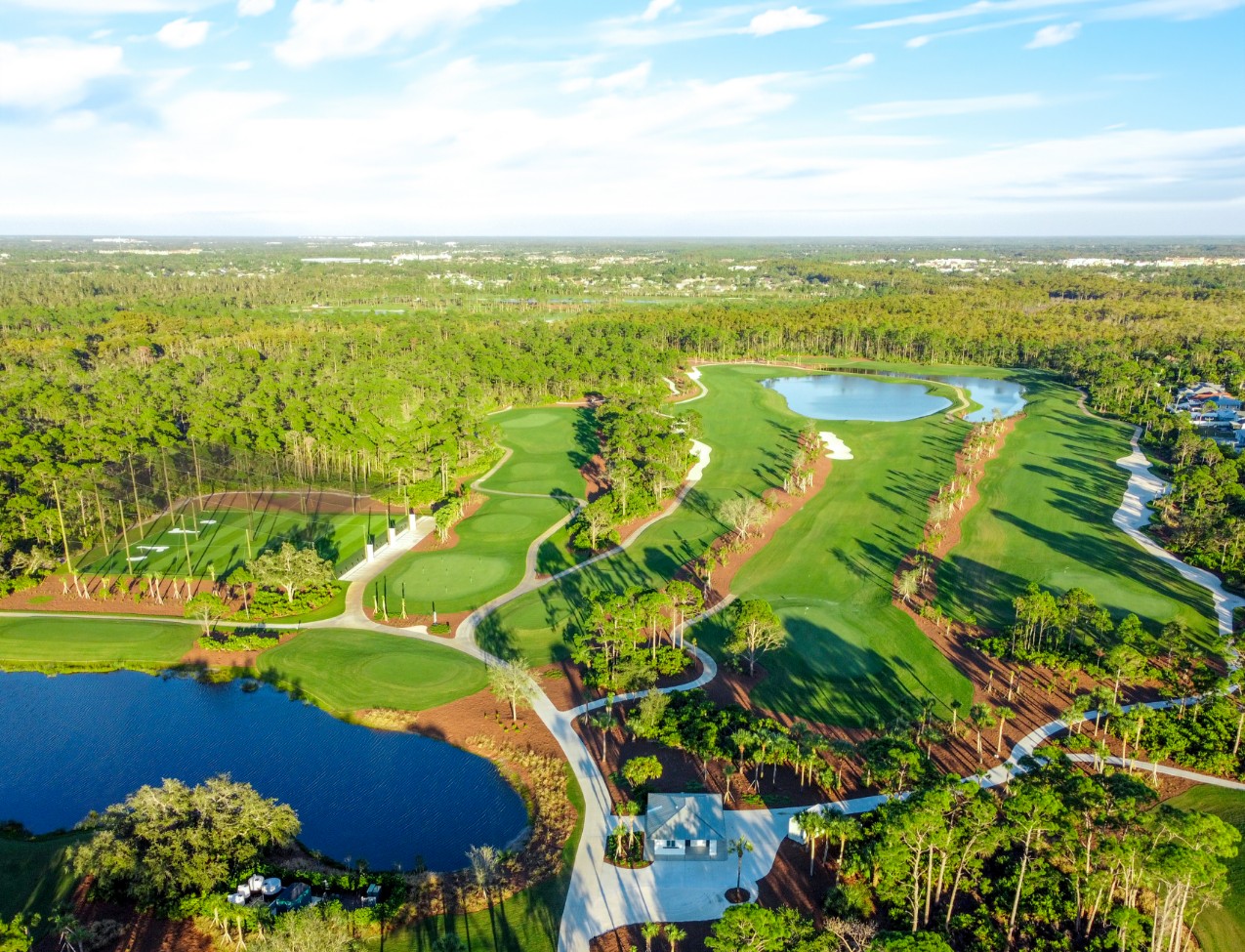 Aerial view of a golf course with green fairways, water hazards, sand traps, and surrounding trees under a partly cloudy sky.