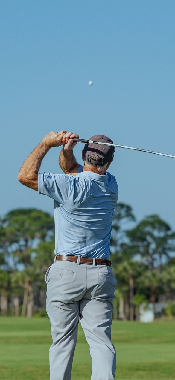 A man wearing a cap and light blue shirt swings a golf club on a golf course, watching the golf ball in the air against a clear blue sky.