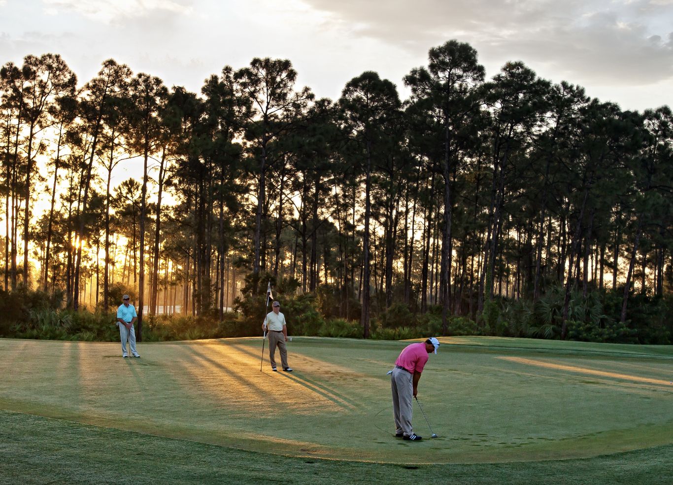 Three people are on a golf course at sunrise, with one person putting while the other two watch, trees silhouetted in the background.