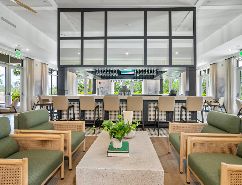 Modern lounge area with green cushioned chairs, a stone coffee table with plants and books, and a bar with beige stools in the background. Large windows provide natural light.