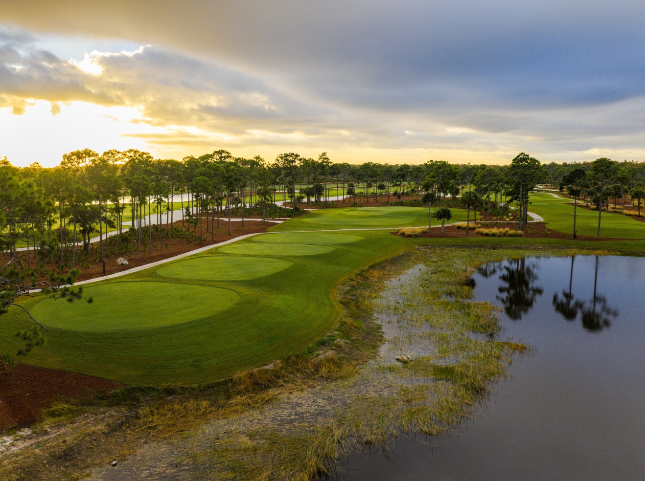 A golf course with neatly mowed greens and fairways sits beside a reflective water hazard, bordered by trees under a partly cloudy sunset sky.