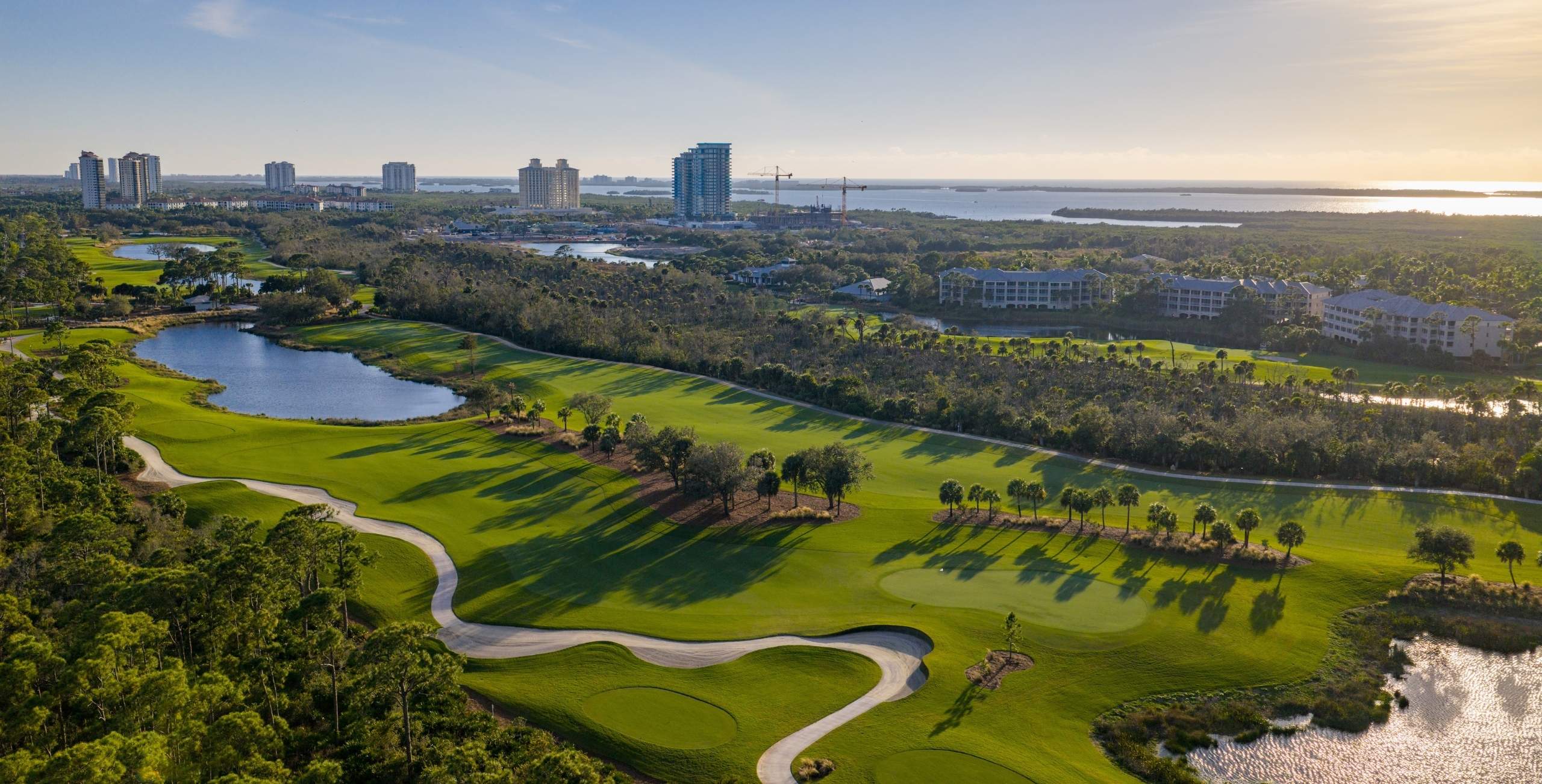 Aerial view of a golf course with sand traps and water features, surrounded by trees, buildings, and a distant body of water under a clear sky.