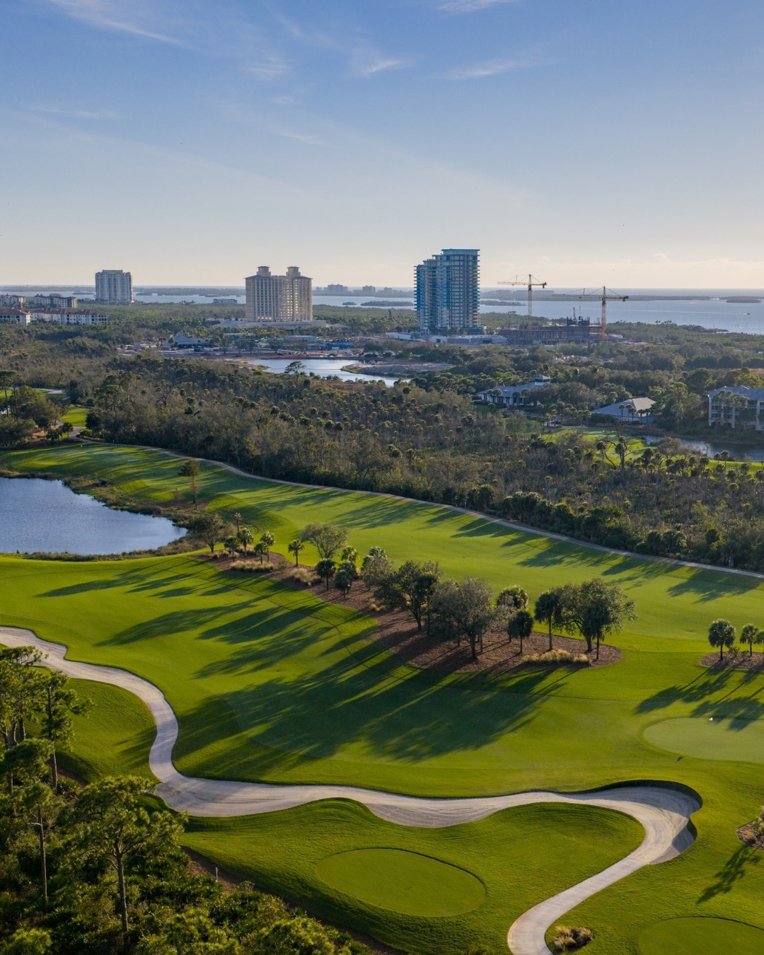 Aerial view of a golf course with green fairways, trees, a water hazard, and nearby city buildings under a clear blue sky.